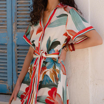 Woman wearing a colorful floral dress leaning against a white wall with blue shutters.