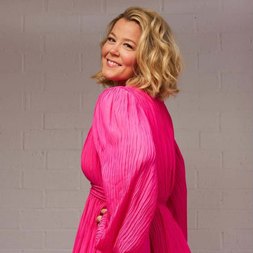 Woman wearing a bright pink pleated dress against a gray brick wall.