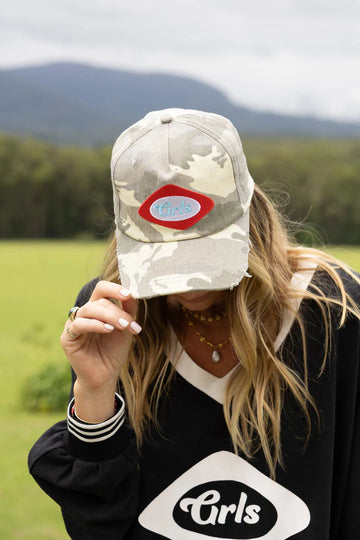 Person wearing a camouflage cap with a logo and a black sweatshirt with 'Girls' text outdoors.
