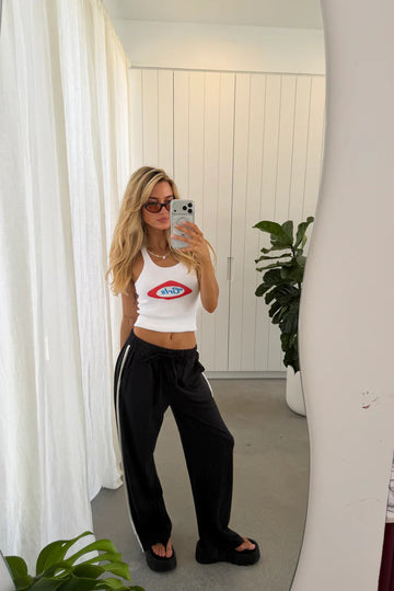 Woman taking a mirror selfie wearing a white tank top with a red and blue design and black pants.