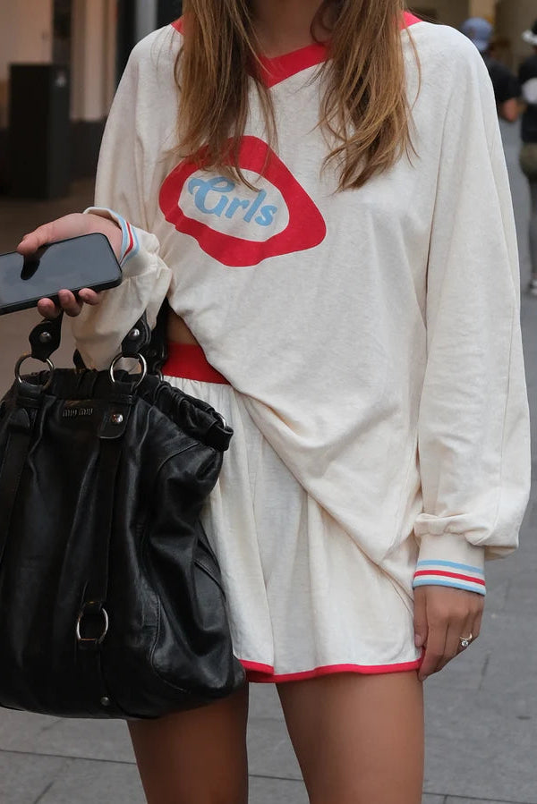 Person wearing a white sweatshirt with a red and blue graphic, holding a black bag and phone.