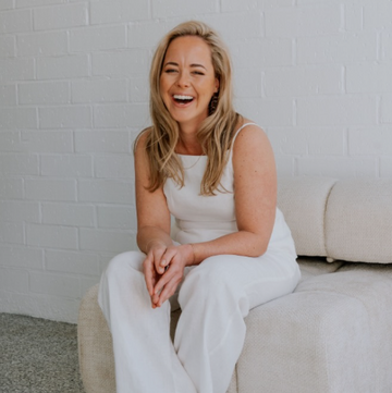 Woman in a white outfit sitting on a beige couch against a white brick wall.