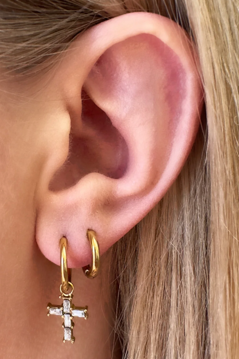 Close-up of an ear wearing gold hoop earrings with a small cross pendant.